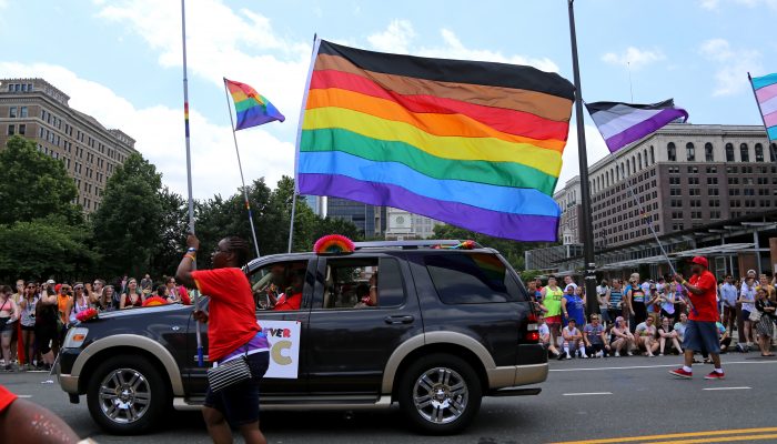 Black car with More Color, More Pride flag attached. People walking in a parade holding Pride flags.