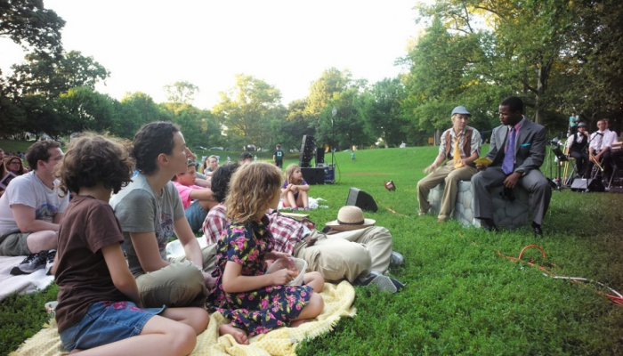 Two men sit on a rock in a park while the crowd watches their performance. 