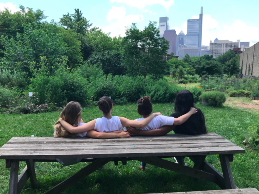 four youth with their backs to the camera sitting at a picnic bench with their arms around each other looking at the skyline of philadelphia