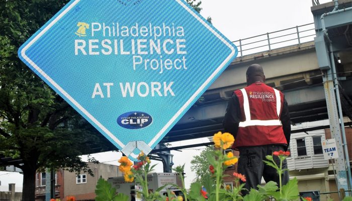 Diamond-shaped street sign that says "Philadelphia Resilience Project at work" with a volunteer standing in the background.
