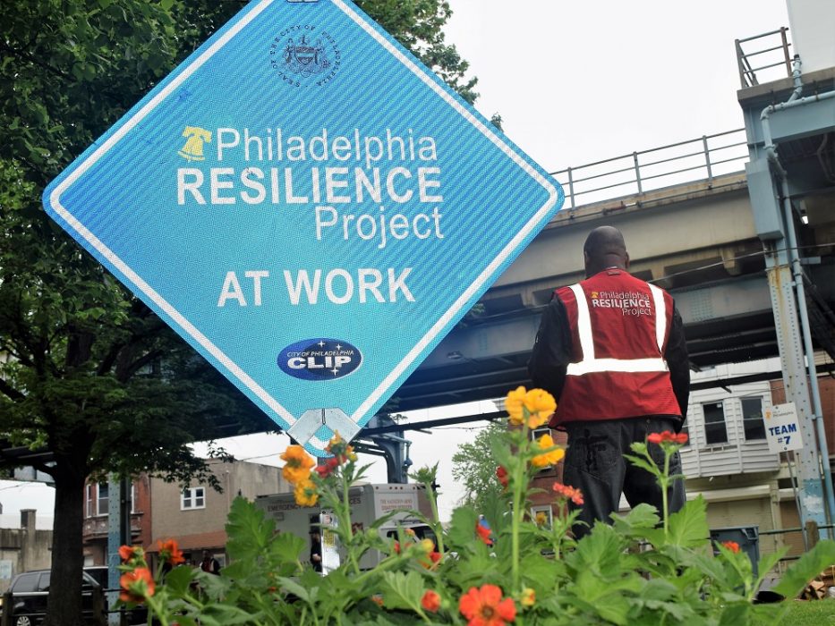 Diamond-shaped street sign that says "Philadelphia Resilience Project at work" with a volunteer standing in the background.