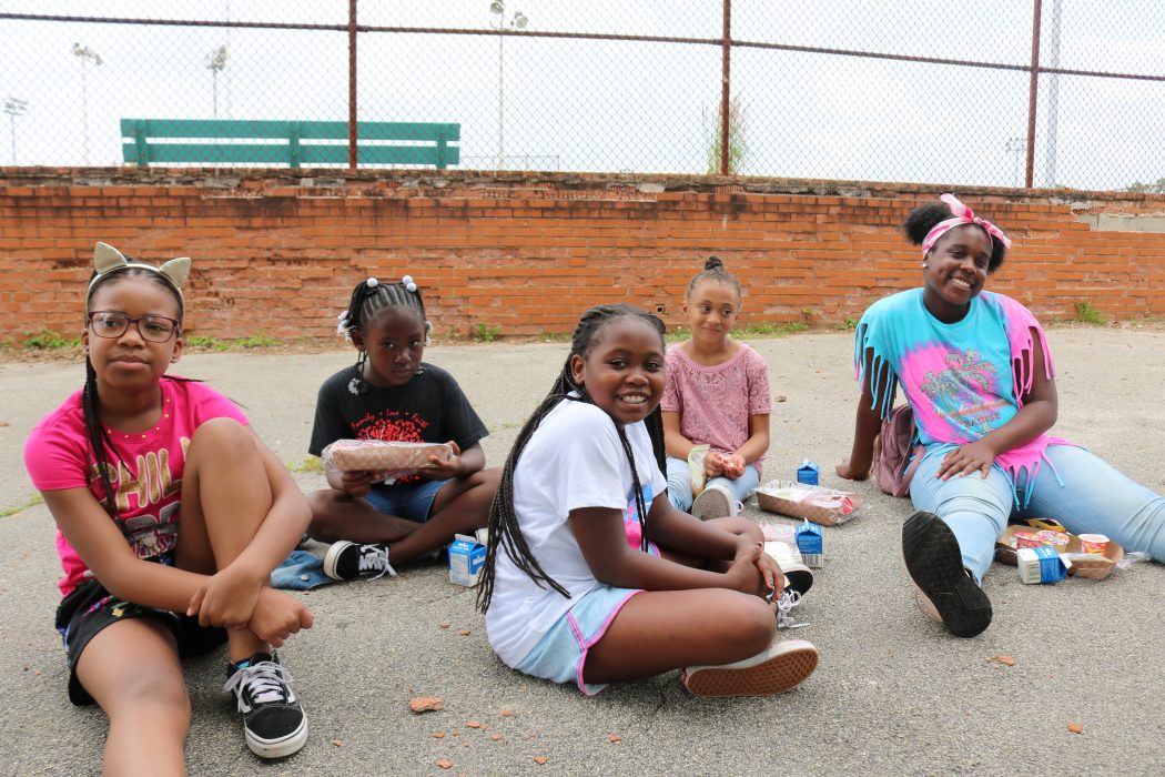 Neighborhood children siting together, smiling, and eating free Summer Meals.