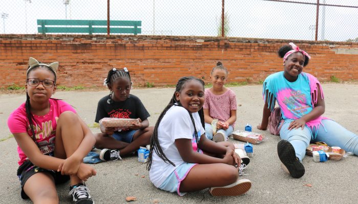 Neighborhood children siting together, smiling, and eating free Summer Meals.