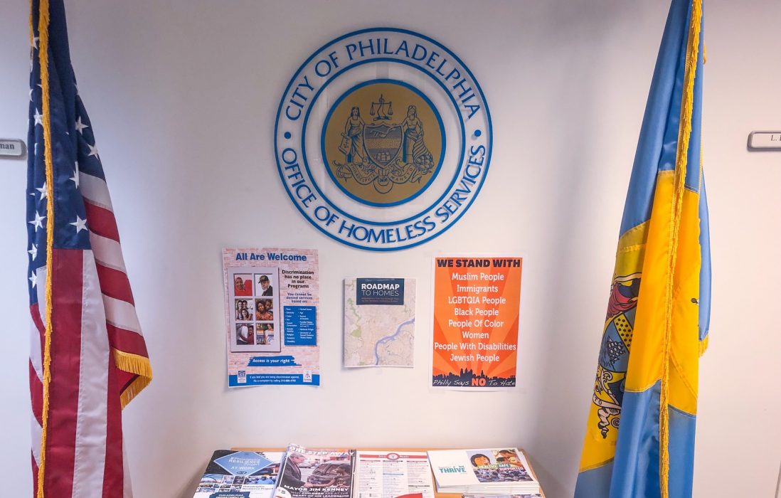 A table with pamphlets and literature below the Office of Homeless Services seal. There is an American flag and a City of Philadelphia flag on either side of the display; one of the signs says, "All are welcome."