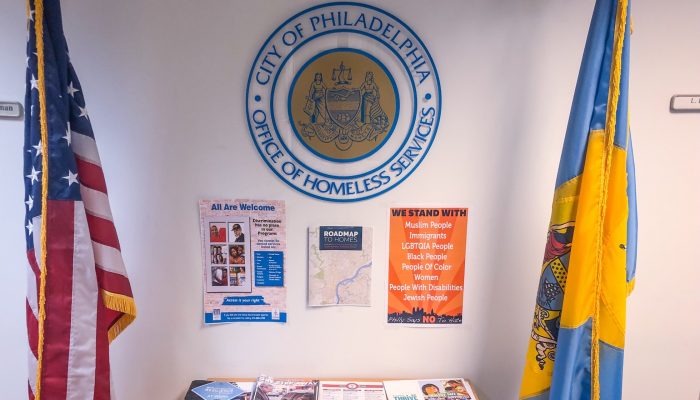 A table with pamphlets and literature below the Office of Homeless Services seal. There is an American flag and a City of Philadelphia flag on either side of the display; one of the signs says, "All are welcome."