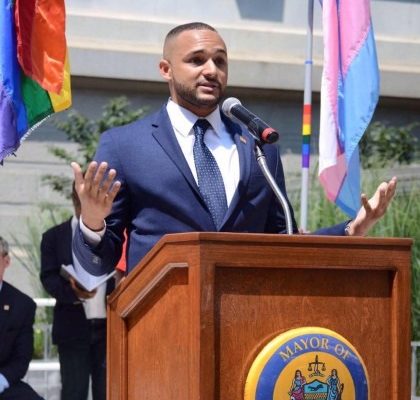 Man at City podium with LGBTQ and Trans Pride flags in background.