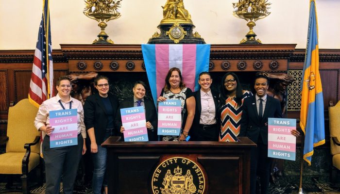 Group poses for photo in front of Trans Pride Flag. 