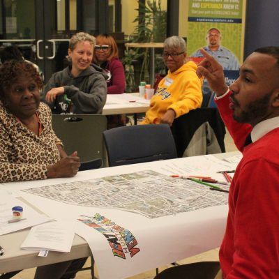 Jose Ferran shares his group's map from the Beat the Heat Design Workshop. There are four people looking on in the background and a map of Hunting Park on the table