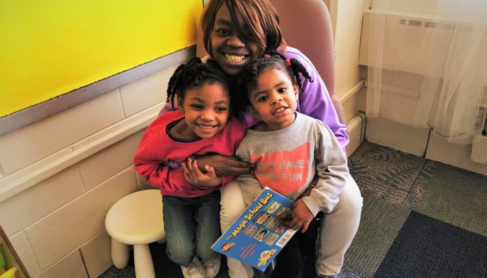 A smiling parent sits in a chair with two children.