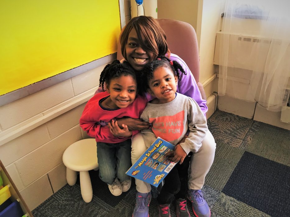 A smiling parent sits in a chair with two children.