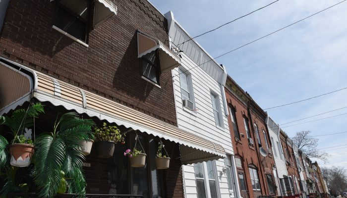 Several two-story brick rowhomes, one with aluminum siding and another with an awning and plants in front of it showing the different way Philly rowhomes, all structured the same, can be customized.