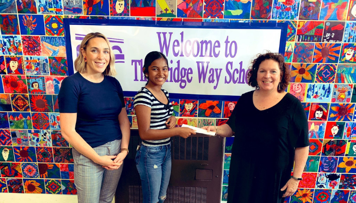 Caitlyn Boyle, Community School Coordinator at George Washington High School, smiles as Daya, a student at George Washington High School, presents the Principle of The Bridge Way School, Rebecca Bonner, with a check in front of a multi-colored wall featured in the hallway of The Bridge Way School.