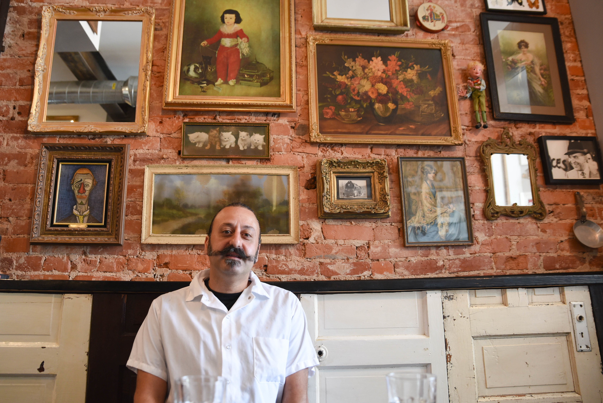 Owner of Stina, Chef Bobby, sits at a table in his restaurant. Behind him is a brick wall decorated with a gallery-style display of artwork. 