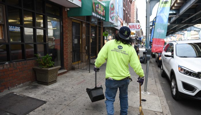 A worker wearing a shirt that reads CLIP and holding a dustbin and broom walking down Kensington Avenue.