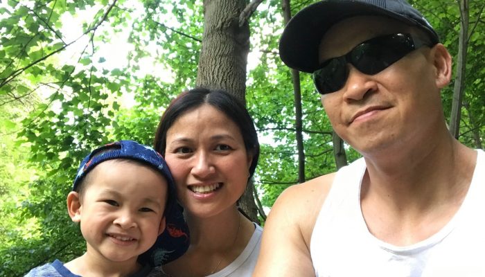 Parents Quang and Ivy smiling with their son Austin in front of green trees.