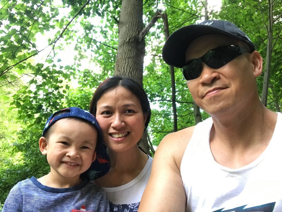 Parents Quang and Ivy smiling with their son Austin in front of green trees.