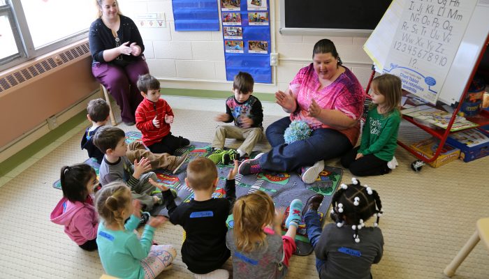 PHLpreK teacher Patty siting on the floor smiling and clapping with students.