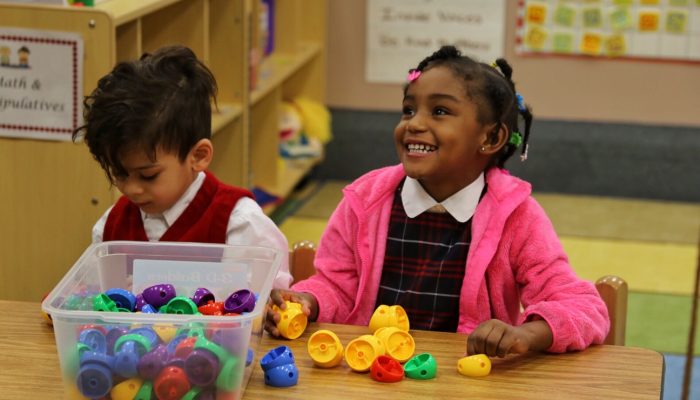 Two PHLpreK students learning together. Colorful learning materials sit on the table in front of them and the student on the right looks up smiling.
