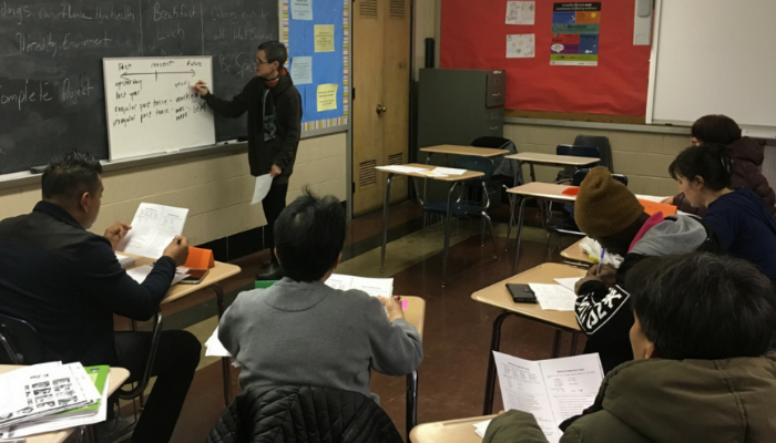 Six adults are sitting in desks inside of a classroom. A teacher is at the front of the classroom, writing a grammar lesson on a whiteboard. The board reads regular past tense and irregular past tense.