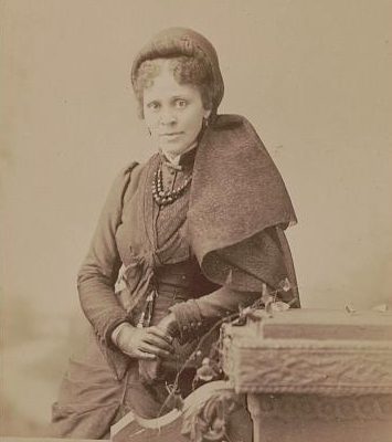 Woman seated with hands in lap, wearing a hat. 