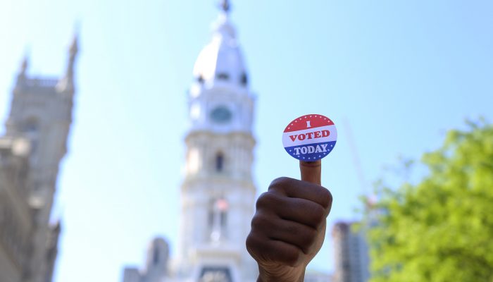Hand holding "I Voted" sticker in front of Philadelphia City Hall.