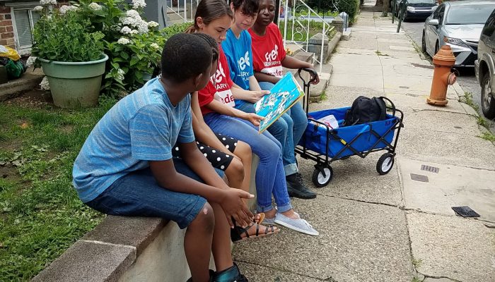 Two children sit outside in front of a house with three adults wearing Free Library of Philadelphia shirts. One adult is reading from a Dr. Seuss book.