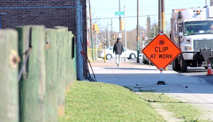 Grass being cut in a vacant lot by workers from CLIP, with a large road sign that says, "CLIP at work" on the sidewalk.