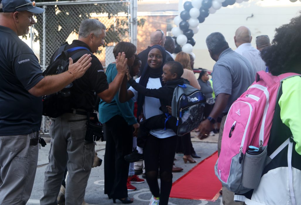 A woman walking through a crowd on a red carpet holds a young boy. Both are high fiving school staff welcoming students back to the first day of school at Robert Morris Elementary.