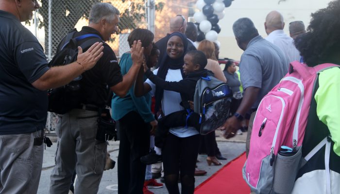 A woman walking through a crowd on a red carpet holds a young boy. Both are high fiving school staff welcoming students back to the first day of school at Robert Morris Elementary.
