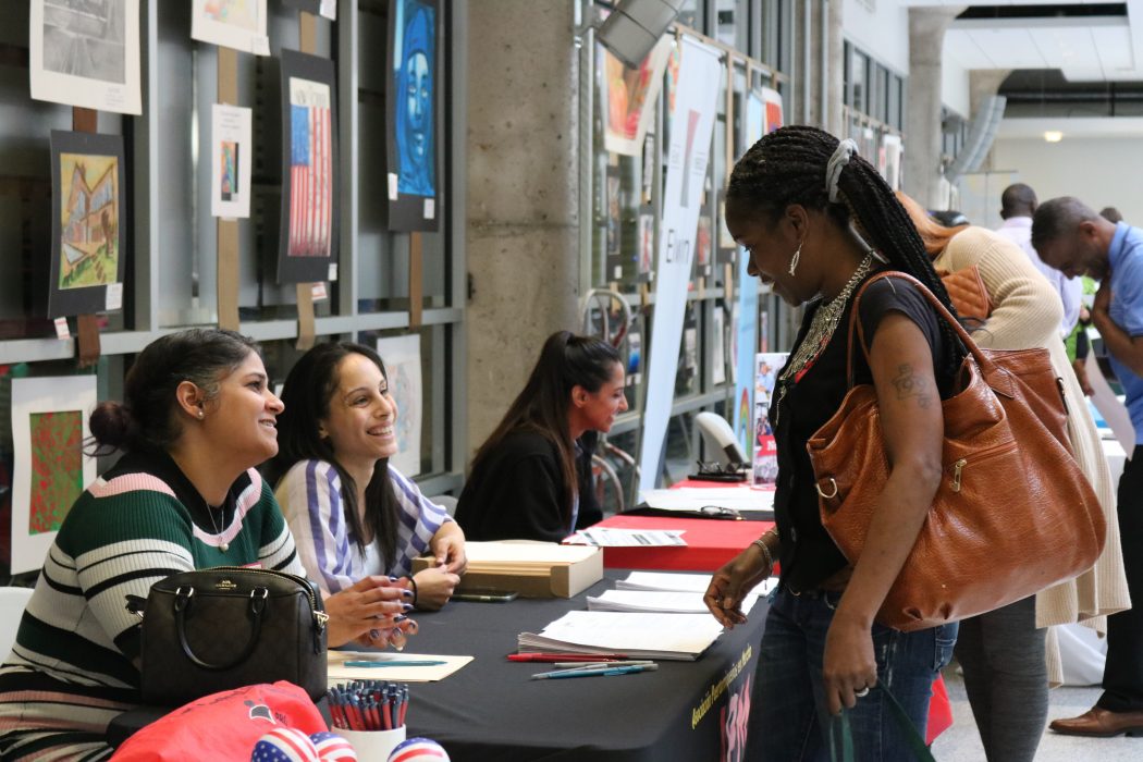 A job seeker looks at an employers table filled with pens and applications. Employees smile at her.