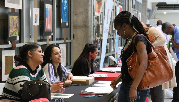 A job seeker looks at an employers table filled with pens and applications. Employees smile at her.