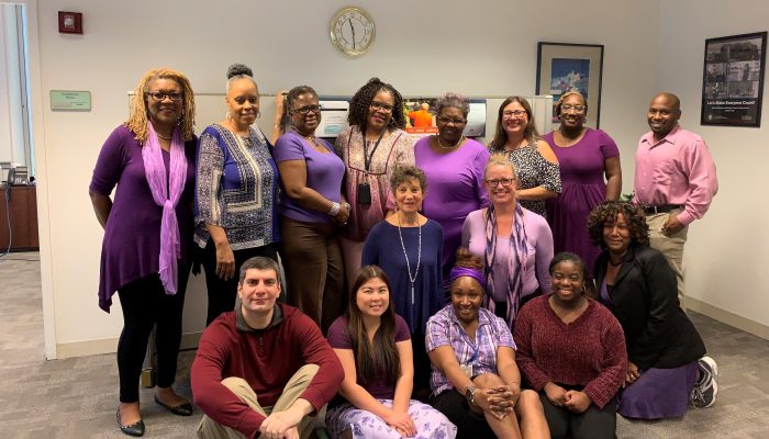A group of OHS employees stand while others kneel, all smiling and wearing purple shirts, scarves, skirts, and dresses to raise awareness of Domestic Violence Awareness Month.