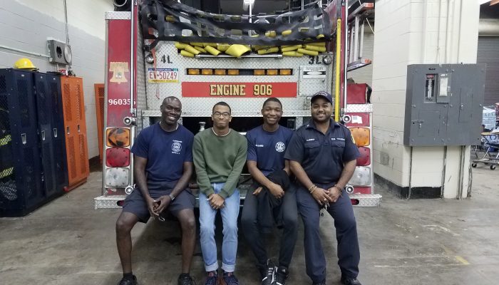 Two adults and two teens sitting on back bumper of fire engine inside garage