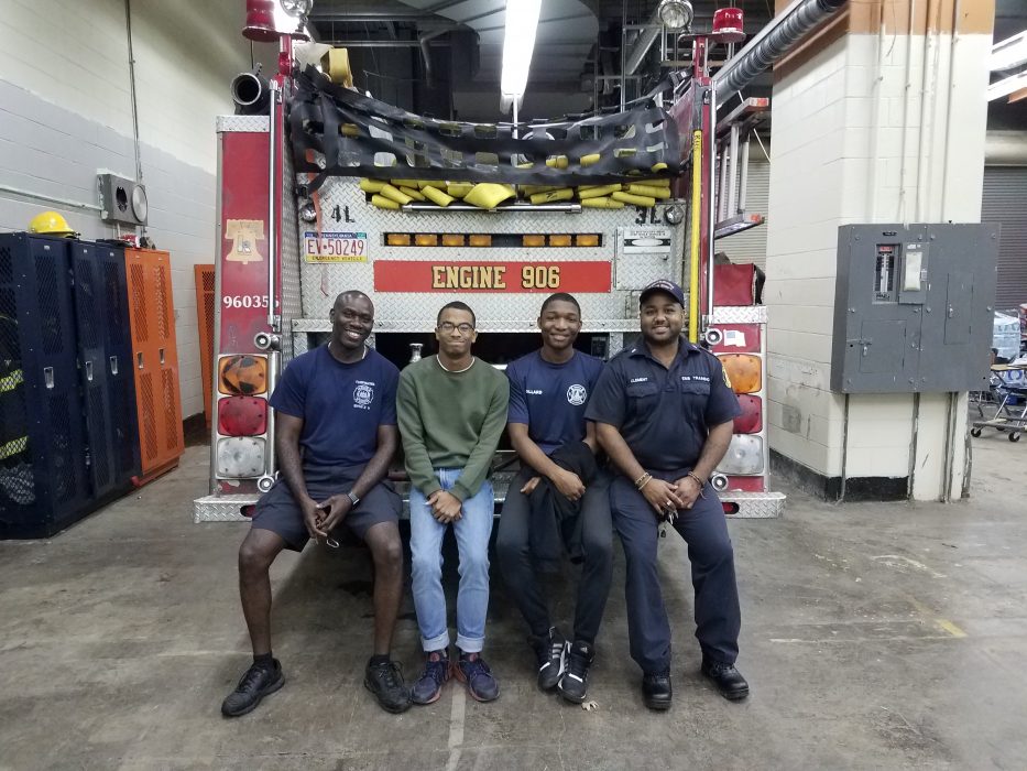 Two adults and two teens sitting on back bumper of fire engine inside garage