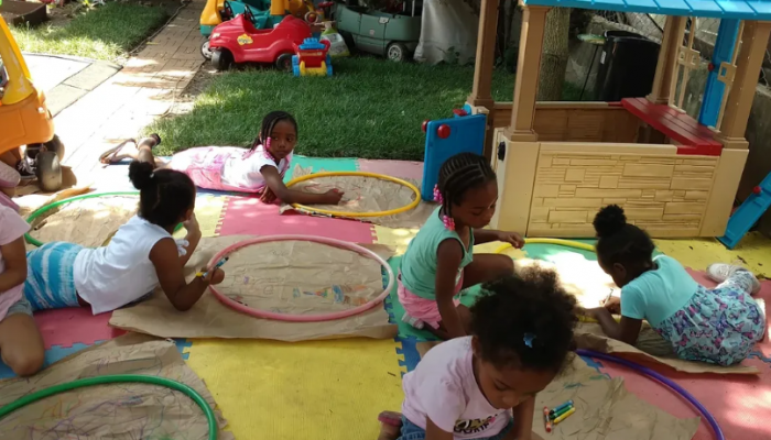 Children sitting and laying outside of Lil' Bits Family Child Care Home. Some are laying on foam squares holding hula hoops. One child is driving a kids car.