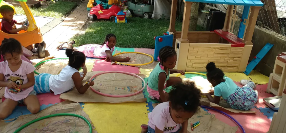 Children sitting and laying outside of Lil' Bits Family Child Care Home. Some are laying on foam squares holding hula hoops. One child is driving a kids car.