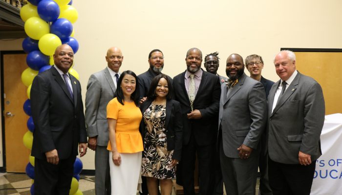 Troy Stratton smiling at the Community School Awards ceremony holding his Community School Award. He is surrounded by City officials. Behind them, balloons and a sign that reads Mayor’s Office of Education decorate the room.