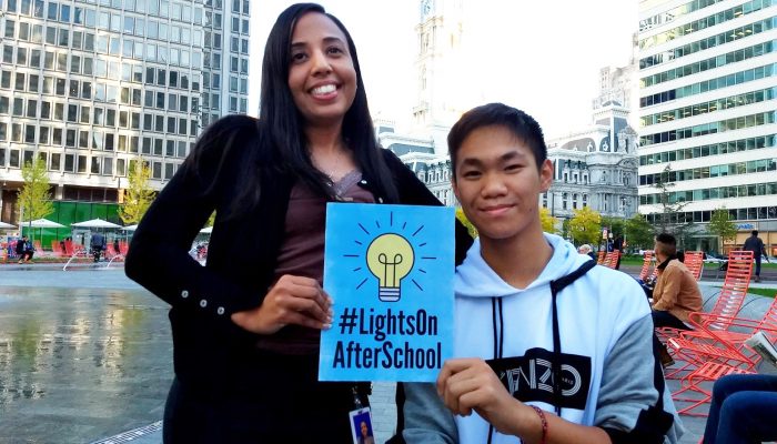 Luz Ayala stands next to Eric Nguyen (seated) in Philadelphia’s LOVE park, both are holding a sign reading #LightsOnAfterSchool.