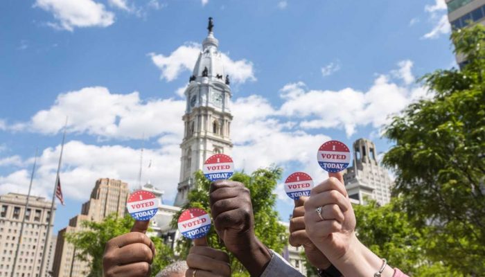 many hands with i voted stickers on their thumbs giving a thumbs up in front of city hall