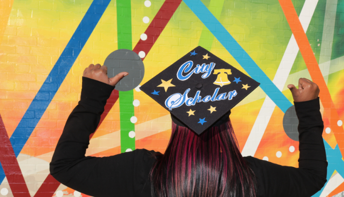 A student with her back turned, points up to a graduation cap that she's wearing. It reads City Scholar. It is decorate with stars, and a City of Philadelphia bell.
