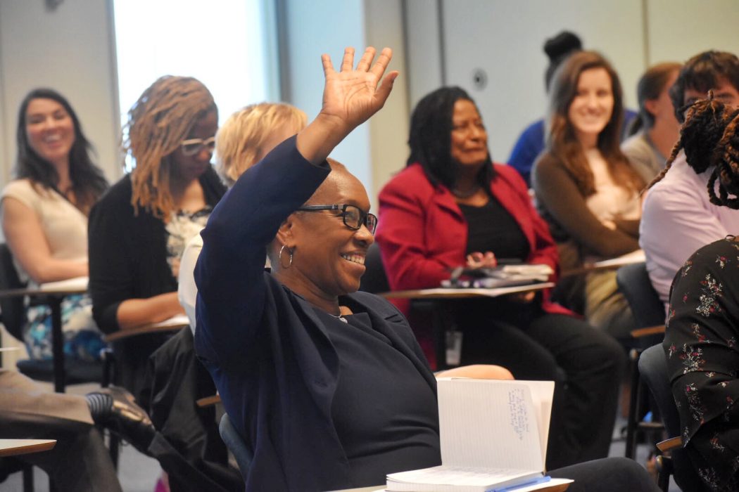 Group of people, each sitting at small desks. Woman at front raises her hand.