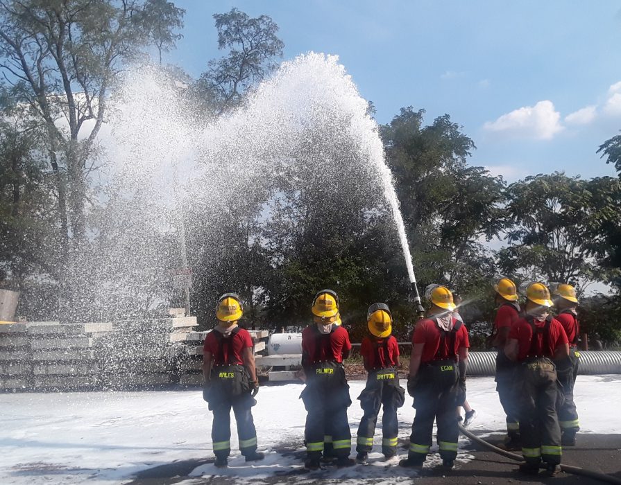 several firefighters watching as hose sprays foam into air outside