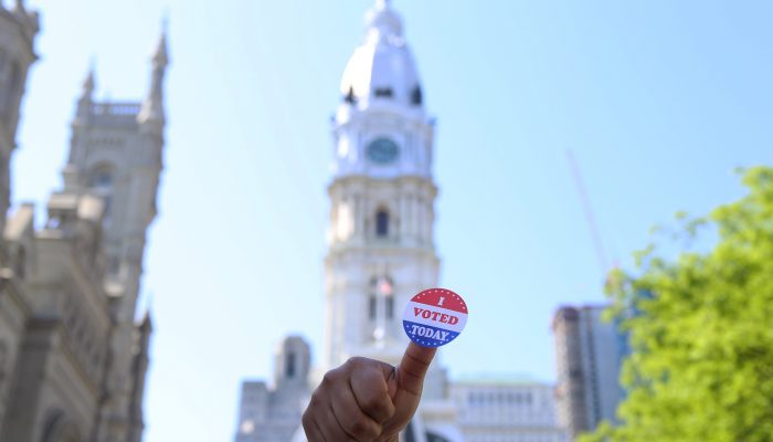 Thumb holding "I Voted" sticker in front of Philadelphia City Hall.