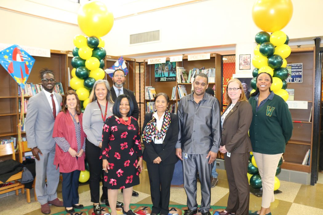 A group of Community School principals stand on stage with Chief Education Officer Otis Hackney and Director of Community School Operations Maxwell Akuamoah-Boateng smiling in front of balloons tower, and bookcases.
