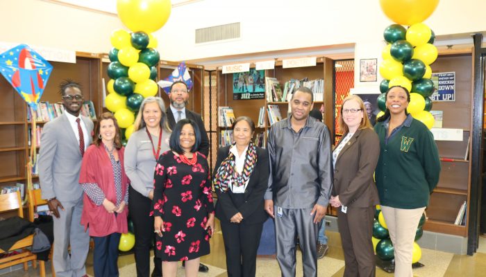 A group of Community School principals stand on stage with Chief Education Officer Otis Hackney and Director of Community School Operations Maxwell Akuamoah-Boateng smiling in front of balloons tower, and bookcases.