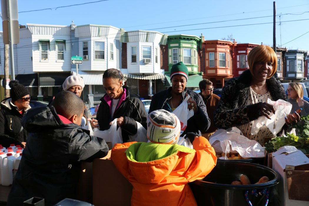 Students pass out fresh produce to community members outside of Edward Gideon School. Kale, potatoes, and bottles of milk are on the table, and Philly row homes are behind them.