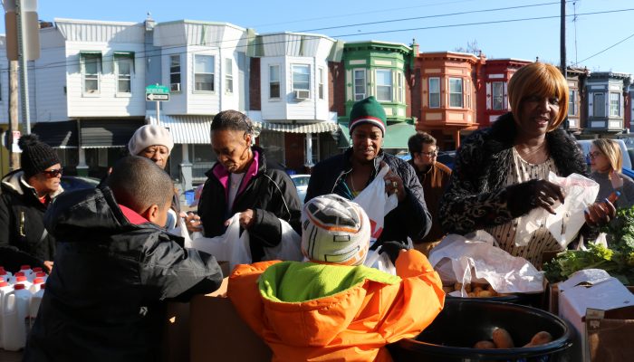 Students pass out fresh produce to community members outside of Edward Gideon School. Kale, potatoes, and bottles of milk are on the table, and Philly row homes are behind them.