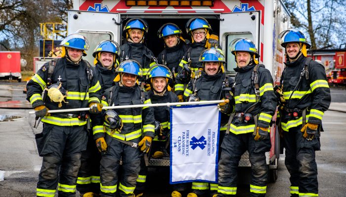 ten paramedics in bunker gear and helmets smiling and standing in back of ambulance.