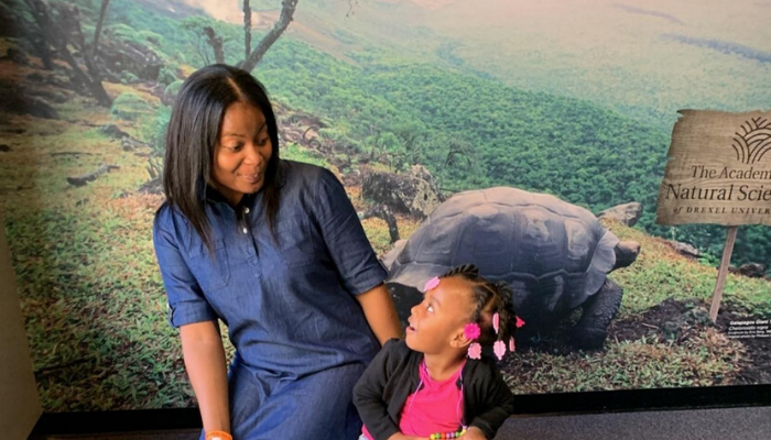 Mother and daughter, Ariel and Alyssa look at each other. Alyssa is excited and her mouth is open. They are in front of a wall that says The Academy of Natural Sciences of Drexel University
