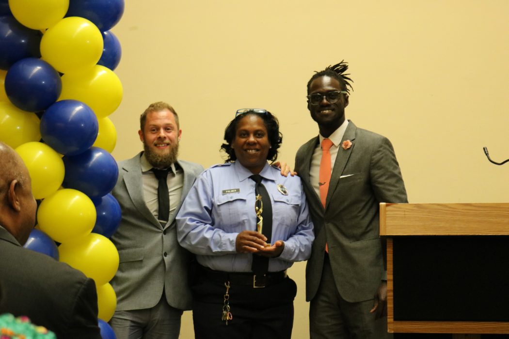 Officer Teresa Palmer, center, standing between William Reed, Community School Coordinator for Cramp Elementary School, and Maxwell Akuamoah-Boateng, Director of Operations for Community Schools, as she holds her Community School champion award trophy. All are smiling.
